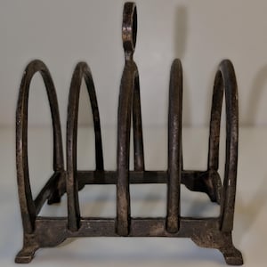 May include: A vintage metal toast rack with a dark, aged finish. The rack features a series of arched dividers and a handle at the top. The base has decorative feet. This kitchenware item is designed to hold multiple slices of toast.