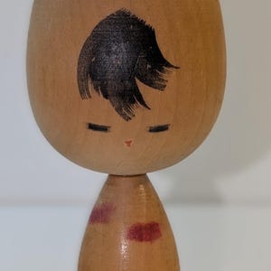 May include: A wooden Kokeshi doll with a rounded head and cylindrical body. The doll features black painted hair, closed eyes, and a small red mouth. The body has red painted accents. The doll is a traditional Japanese craft.
