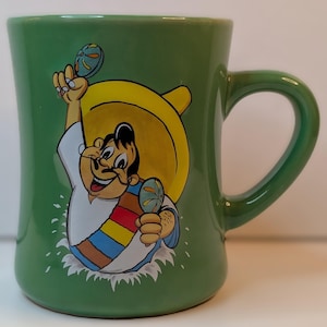 May include: Green ceramic coffee mug with a cartoon illustration of a mustached man wearing a sombrero and holding maracas. The mug has a curved handle and a wide base. The illustration is in bright colors.