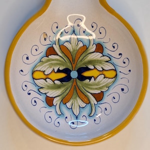 May include: A white ceramic spoon rest with a yellow rim and handle. The center features a symmetrical floral design in green, yellow, brown, and blue. Decorative blue swirls surround the central design, creating a classic, elegant look.
