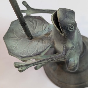 May include: A bronze frog figurine with an open mouth, perched on a lily pad. The frog is detailed with textured skin and outstretched limbs. A dark rod extends from the lily pad. The base is a circular shape.