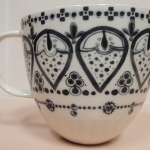 May include: White ceramic mug with a large handle, decorated with a repeating black and gray pattern. The design features stylized heart shapes filled with dots and floral elements, creating a decorative and elegant look.