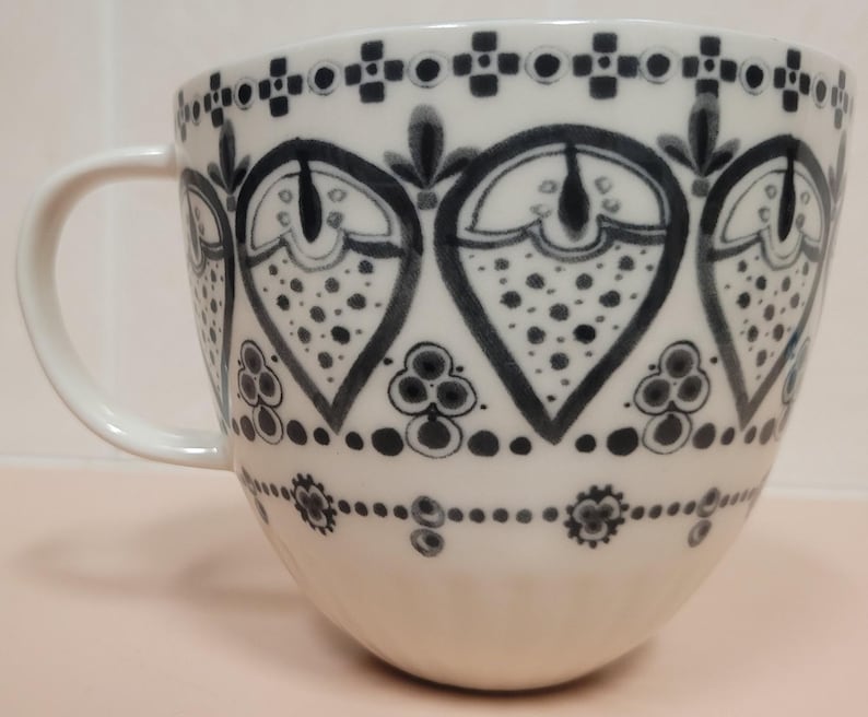 May include: White ceramic mug with a large handle, decorated with a repeating black and gray pattern. The design features stylized heart shapes filled with dots and floral elements, creating a decorative and elegant look.