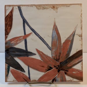 May include: Square ceramic tile with a floral design. The artwork features large, stylized leaves in shades of red, black, and gray against a cream-colored background. A dark blue line intersects the design. The tile has a distressed, aged appearance.