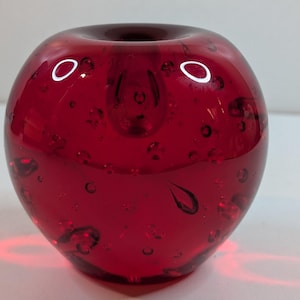 May include: A round, red glass vase with a small opening at the top. The vase is filled with small air bubbles, giving it a textured appearance. The red glass is translucent, allowing light to pass through.