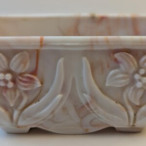 May include: A square, off-white ceramic bowl with a floral design. The bowl features embossed flowers and leaves on the sides, with a marbled effect of pink and orange hues. The bowl is approximately 4 inches wide and 2 inches tall.