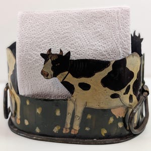 May include: A metal napkin holder with a cow design. The holder is painted black and features a cow with black and white spots. The cow is standing in front of a stack of white paper napkins. The holder has a handle on each side.
