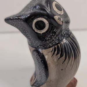 May include: A hand-painted ceramic owl figurine. The owl has a black body with white dotted accents, large white-rimmed eyes, and a gray chest. The feet are brown, and the beak is black. The owl is in a seated position.