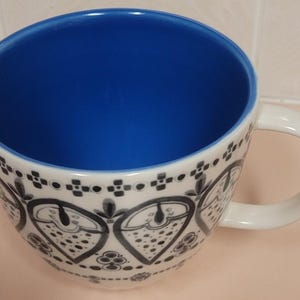 May include: White ceramic coffee mug with a blue interior and a decorative black and white pattern around the exterior. The mug has a large, rounded shape and a curved handle.