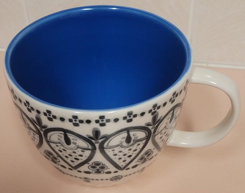 May include: White ceramic coffee mug with a blue interior and a decorative black and white pattern around the exterior. The mug has a large, rounded shape and a curved handle.