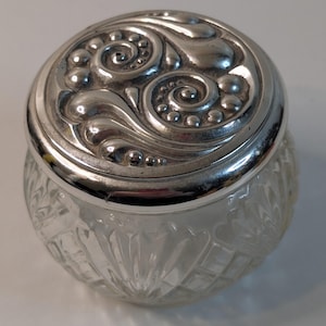 May include: A clear glass jar with a silver lid. The lid has an ornate design with swirling patterns and small, raised dots. The jar's body has a diamond-like pattern. The jar is likely for storing small items.