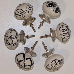 May include: Six ceramic cabinet knobs with floral and geometric black designs on a cream background. Each knob has a silver-toned base and a threaded bolt for installation. The knobs are arranged in a circular pattern.