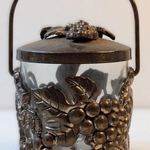 May include: A clear glass ice bucket with a metal lid and handle. The bucket is decorated with a silver-colored grape and leaf design. The handle is arched and attached to the sides of the bucket.
