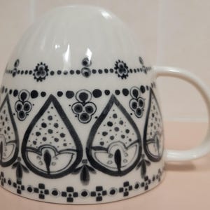 May include: White ceramic mug with a curved handle, decorated with a repeating black and gray floral pattern. The design features teardrop shapes filled with dots and stylized flowers, creating a decorative and elegant look. The mug is suitable for hot beverages.