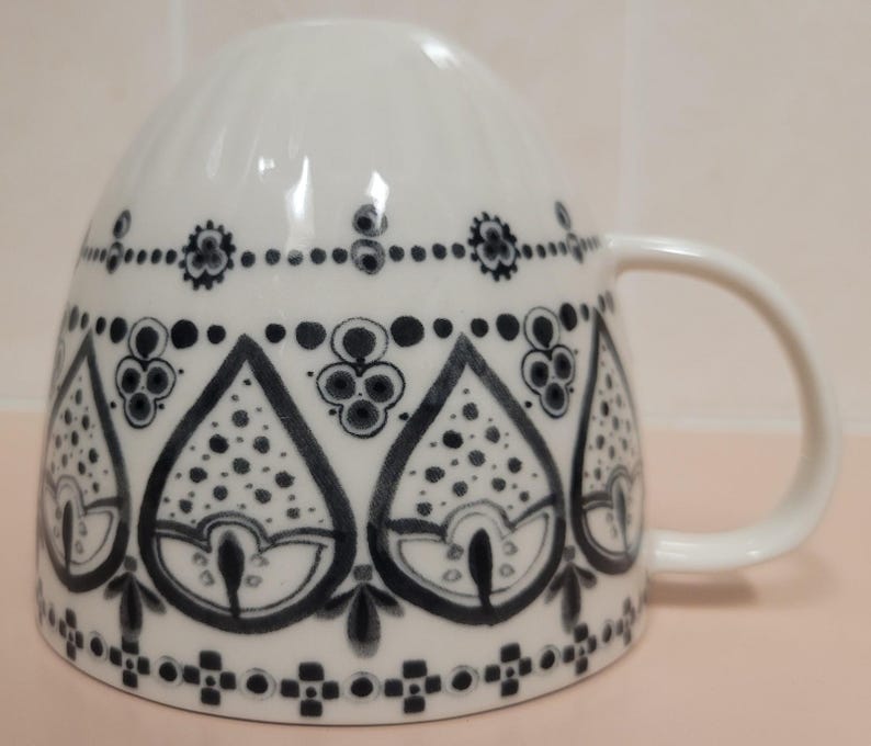 May include: White ceramic mug with a curved handle, decorated with a repeating black and gray floral pattern. The design features teardrop shapes filled with dots and stylized flowers, creating a decorative and elegant look. The mug is suitable for hot beverages.