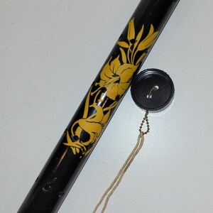 May include: A black cylindrical container with a screw-on cap and a decorative yellow floral design. The cap is attached to the container with a small chain and a beige string. The container has several small holes.