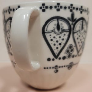 May include: White ceramic mug with a large, curved handle. The mug is decorated with a repeating pattern of black heart-shaped designs and dotted accents. The design is repeated around the rim and base of the mug.
