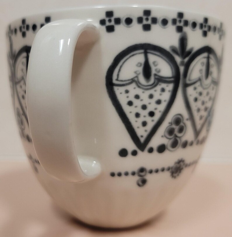 May include: White ceramic mug with a large, curved handle. The mug is decorated with a repeating pattern of black heart-shaped designs and dotted accents. The design is repeated around the rim and base of the mug.