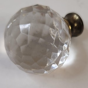May include: A clear, faceted crystal cabinet knob with a brass-colored base. The knob is spherical, with numerous flat surfaces creating a geometric pattern. The brass base is circular and slightly domed.