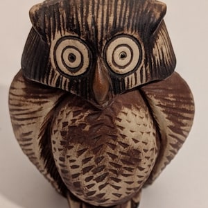 May include: A ceramic owl figurine with a detailed, textured surface. The owl features large, concentric circle eyes, a brown beak, and a mix of brown and beige tones. The wings and chest have carved patterns.