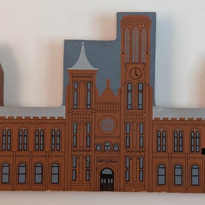 May include: A wooden, hand-painted depiction of a building, likely a museum or government structure. The building is primarily brown with gray and blue accents. The word "Smithsonian" is visible above the entrance. The clock tower has a clock face.