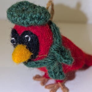 May include: A handmade, felt bird ornament with a red body, black face, and yellow beak. The bird wears a green knitted hat and scarf. The bird's feet are brown, and a string is attached for hanging. A festive decoration.