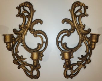 May include: Pair of ornate, gold-colored wall sconces. Each sconce features a decorative, swirling design with floral accents and two candle holders. The sconces are made of a metallic material and are designed to be mounted on a wall.