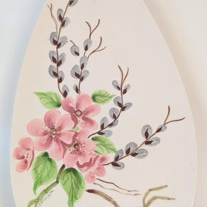 May include: Cream-colored, egg-shaped decorative plate with a floral design. Pink flowers with green leaves and gray pussy willows are painted on the plate. The plate has a smooth surface and a slightly rounded shape.