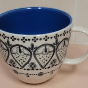 May include: White ceramic coffee mug with a blue interior and a white handle. The exterior features a black and white floral pattern. The mug is designed with a wide, rounded shape.
