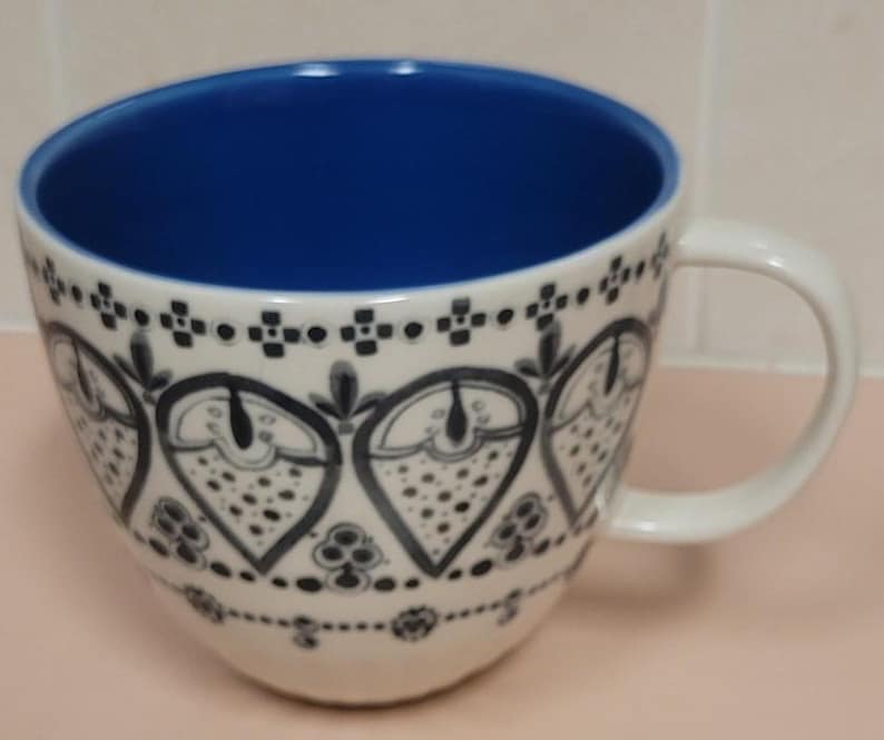 May include: White ceramic coffee mug with a blue interior and a white handle. The exterior features a black and white floral pattern. The mug is designed with a wide, rounded shape.