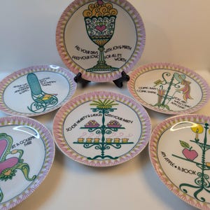 May include: A set of six decorative plates with whimsical designs and text. The plates feature a pink and white border and various illustrations, including chairs, a goblet, and a tiered dessert stand. The text includes phrases like "Fill your days with joy" and "So eat hearty & laugh."