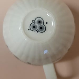 May include: A white teacup with a ribbed exterior and a small handle. The bottom of the cup features a black and gray floral design. The cup is made of ceramic and is suitable for hot beverages.