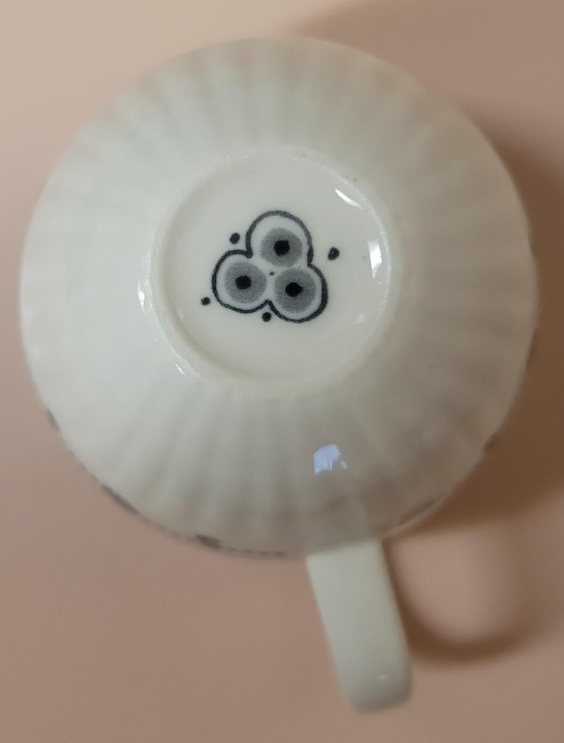 May include: A white teacup with a ribbed exterior and a small handle. The bottom of the cup features a black and gray floral design. The cup is made of ceramic and is suitable for hot beverages.