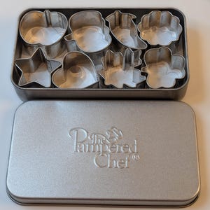 May include: A set of eight metal cookie cutters in various shapes, including a star, heart, and flower, arranged in a rectangular tin. The tin is silver and has the words "The Pampered Chef" embossed on the lid.