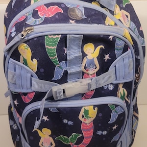 May include: A navy blue backpack featuring a repeating pattern of mermaids with yellow, pink, and green tails. The backpack has light blue accents, straps, and a zipper closure. It includes a top handle and adjustable shoulder straps.