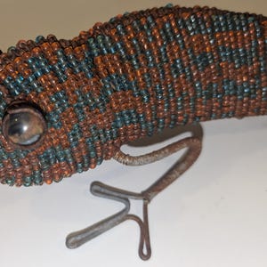 May include: A beaded lizard sculpture in shades of brown and teal. The lizard's body is covered in small beads, with a detailed eye and metal legs. The sculpture is a decorative item.