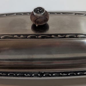 May include: Silver-toned butter dish with a lid and a decorative border. The lid features a wooden knob and a rectangular design. The dish is oval-shaped, with a black and silver patterned border. A classic kitchenware item.