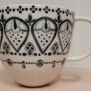 May include: A white ceramic mug with a large handle, decorated with a repeating pattern of dark blue heart-shaped designs filled with dots. The rim and base feature additional dark blue geometric patterns.