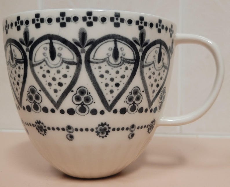 May include: A white ceramic mug with a large handle, decorated with a repeating pattern of dark blue heart-shaped designs filled with dots. The rim and base feature additional dark blue geometric patterns.