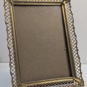 May include: A rectangular brass picture frame featuring an intricate lattice or filigree border design. The frame has a metallic gold-toned finish with decorative corner accents and a plain, neutral-colored backing insert visible inside.