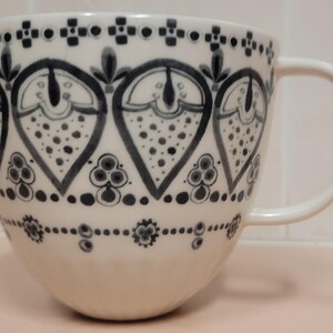May include: White ceramic mug with a large handle, decorated with a repeating pattern of stylized floral and geometric shapes in shades of gray and black. The mug has a wide, rounded shape, suitable for coffee or tea.