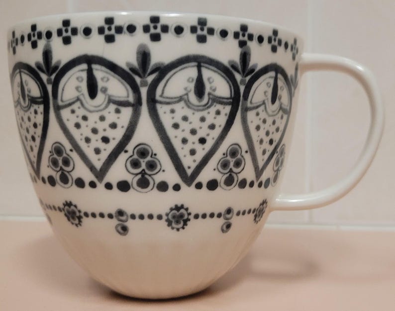 May include: White ceramic mug with a large handle, decorated with a repeating pattern of stylized floral and geometric shapes in shades of gray and black. The mug has a wide, rounded shape, suitable for coffee or tea.