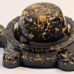 May include: Black turtle-shaped decorative container with gold leaf accents. The container has a round top with the text "THE GOLDEN TOWER COLUMBUS" and a small gold button. The turtle has four feet and a circular opening.