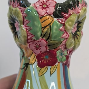 May include: A ceramic vase featuring a flared rim with three-dimensional sculpted flowers in shades of pink, red, and green. The body of the vase displays a light blue background with vertical stripes in various colors including yellow, red, and green.