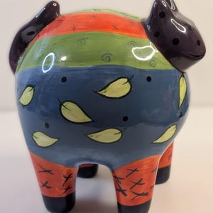 May include: A whimsical ceramic piggy bank with a colorful design. The bank features a dark purple head and tail, with a body painted in horizontal stripes of red, green, and blue. Yellow leaf and black dot accents decorate the blue section. The legs are red with black accents.