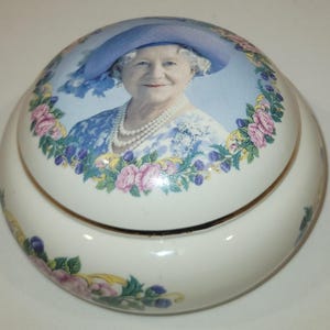 May include: A white ceramic trinket box with a round lid featuring a portrait of Queen Elizabeth II wearing a blue hat and a pearl necklace. The lid is decorated with a floral border of pink and white roses, green leaves, and purple flowers.