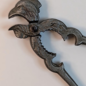 May include: A vintage metal nutcracker with a rooster head design. The nutcracker is silver-colored with intricate details, including a textured finish and decorative curves. The rooster's beak forms the cracking mechanism.
