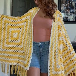 Peut inclure: Un châle au crochet jaune et blanc fait à la main, avec un motif carré et des franges. Le châle est porté sur un haut pêche et un short en jean. Les dimensions du châle sont d'environ 152 cm sur 152 cm.