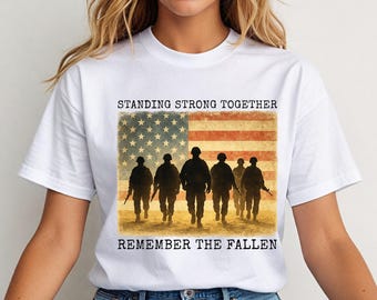 May include: White t-shirt featuring a graphic of silhouetted soldiers walking in front of an American flag. The text "STANDING STRONG TOGETHER" and "REMEMBER THE FALLEN" are printed above and below the image.