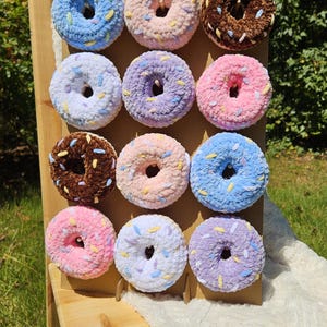 May include: A display of twelve plush donuts in various pastel colors, including blue, pink, purple, and brown. Each donut is decorated with colorful sprinkles and arranged on a wooden stand, creating a playful and inviting aesthetic.